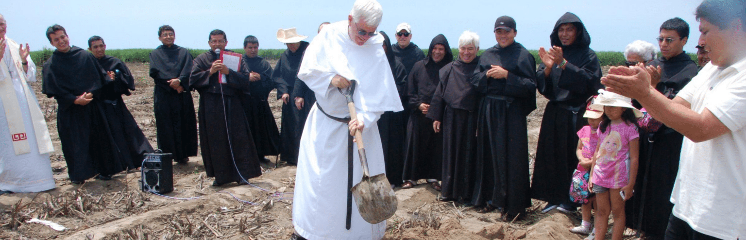 Friar on a foreign mission holding a shovel, digging into the ground, with a group of other friars clapping around him.